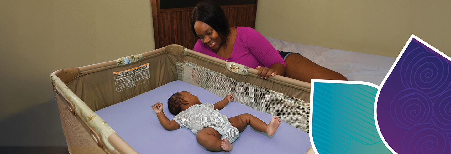 A mother sitting on her bed smiles and leans over to look at her baby lying on his back in an empty crib.