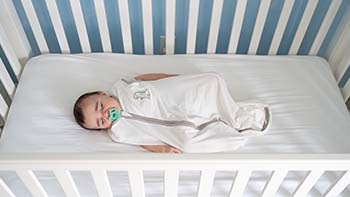 A baby wearing a sleep sack with a pacifier in his mouth sleeping on his back in an empty crib.