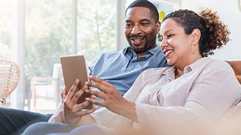 A man and woman seated on a couch, both focused on a tablet in their hands.